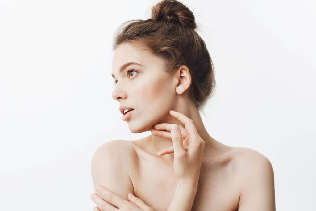 Close up portrait of young female brunette student girl with bun hairstyle and baked shoulders looking aside with calm face expression touching jawline with fingers. Beauty and health.